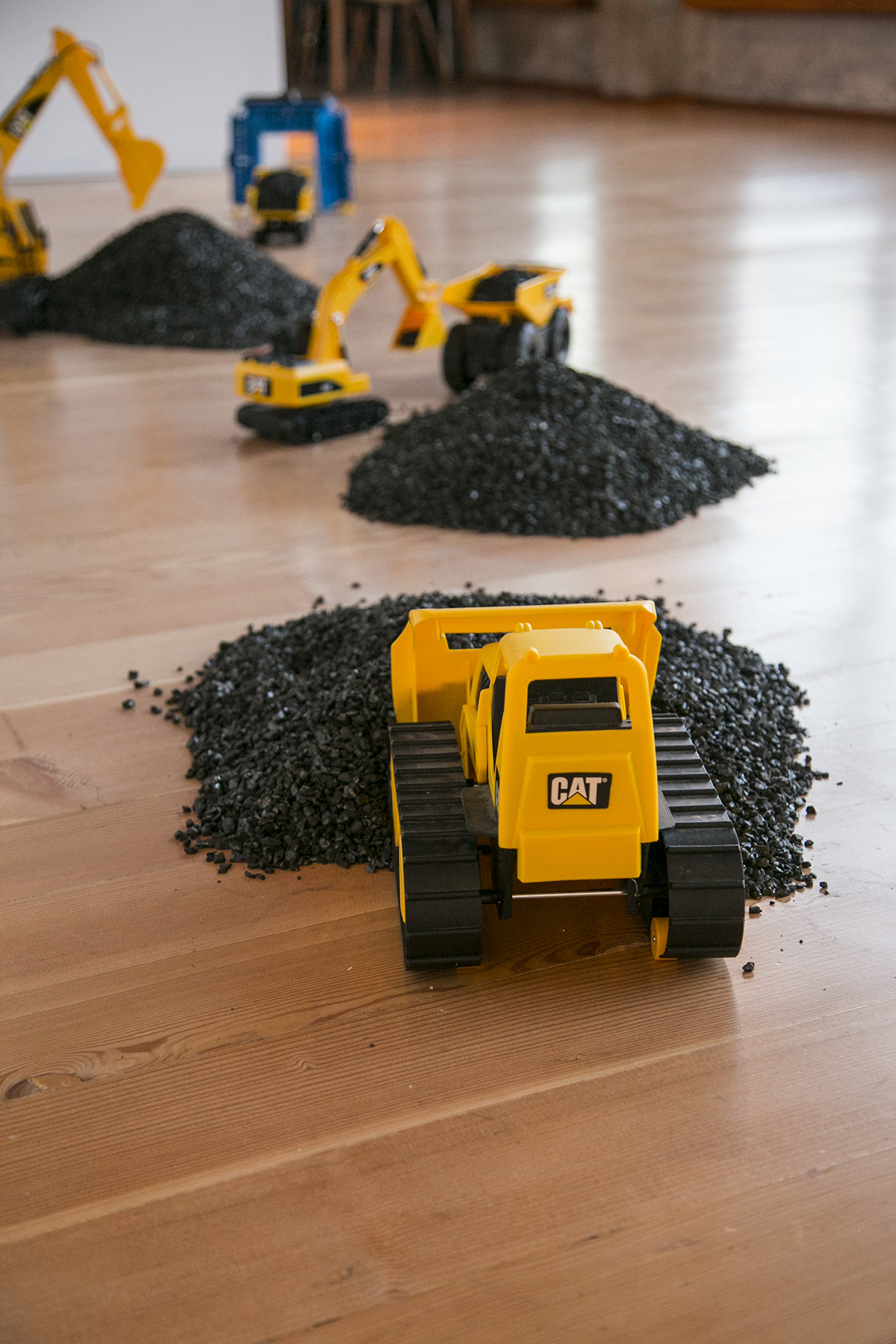 Close-up image of artwork by RYAN! Feddersen includes yellow toy trucks, coal, and blue scrub brush arch