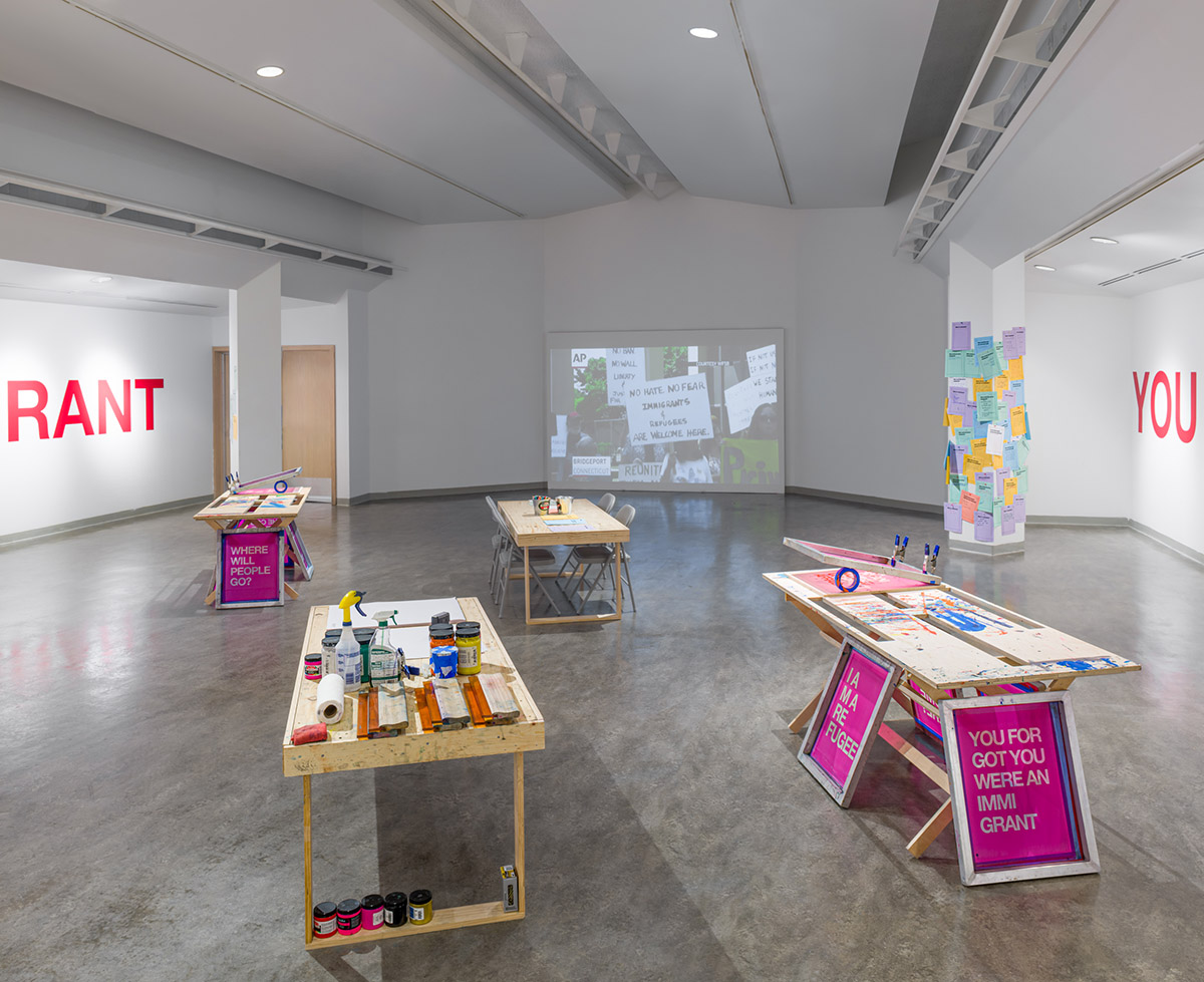 Image of tables set up for screenprinting in the foreground, a large video projection showing protest images in the back. 