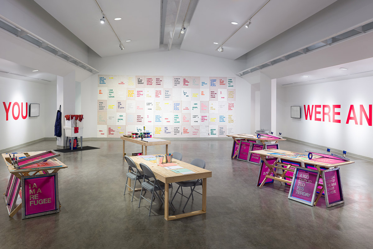 screenprint workshop tables set up to print in the foreground, large wall covered in a grid of printed posters in the back