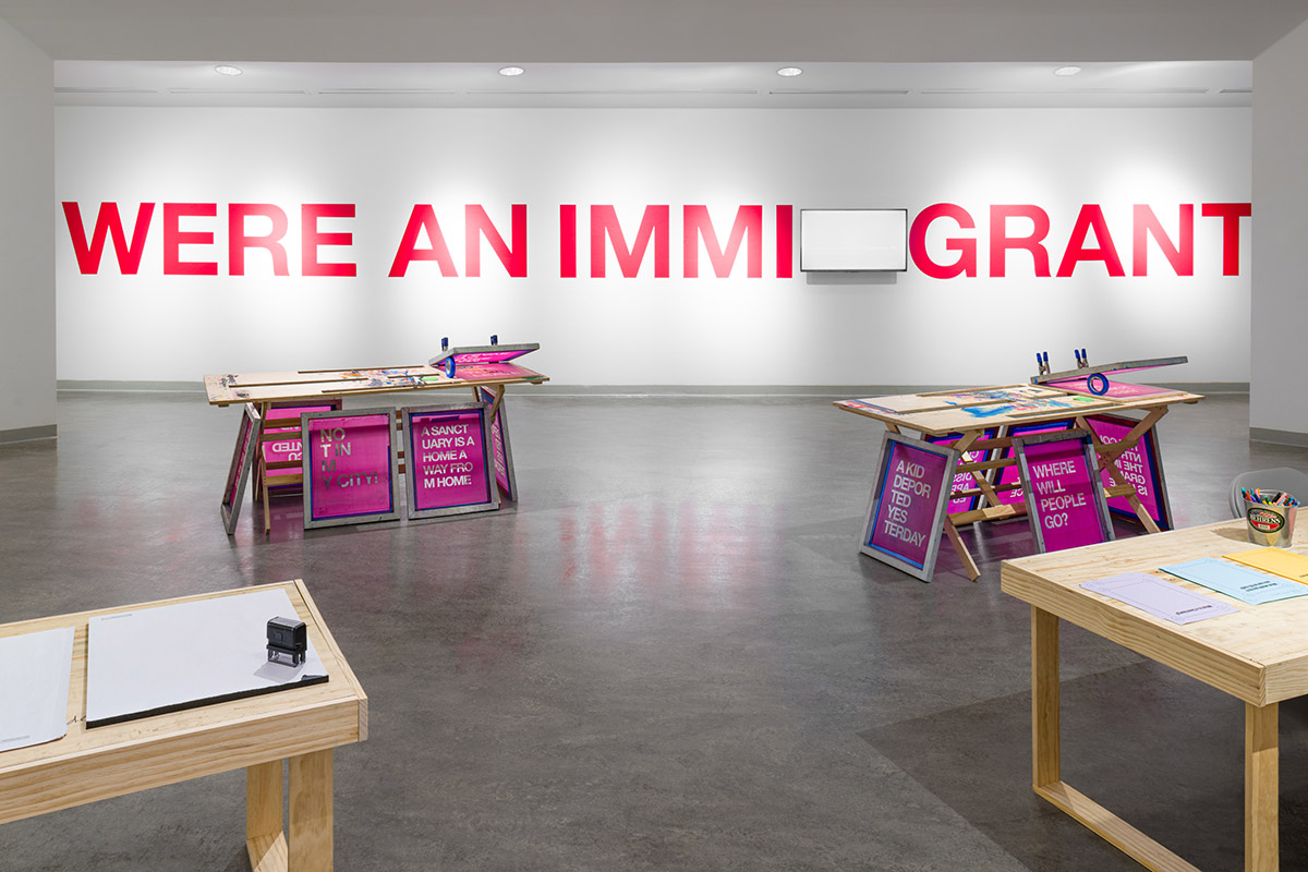 tables set up for screenprint workshop, with video monitor and large red text on a white wall behind the tables