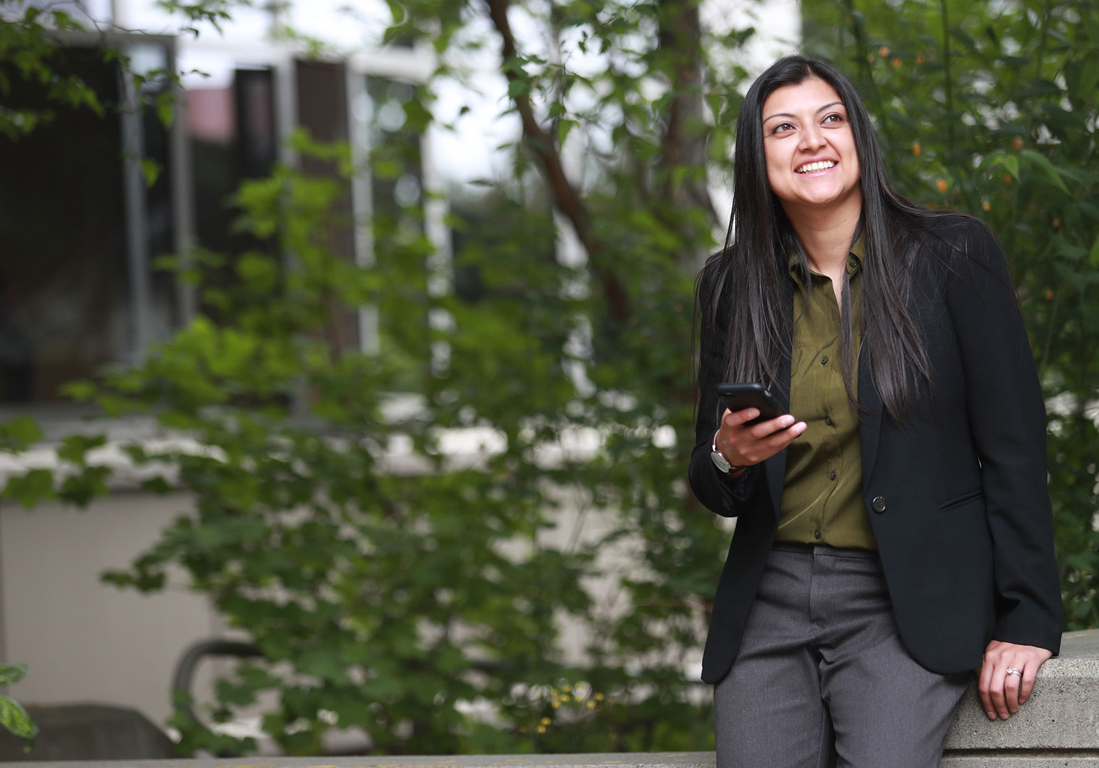 Angela standing in front of greenery while smiling and holding her phone.