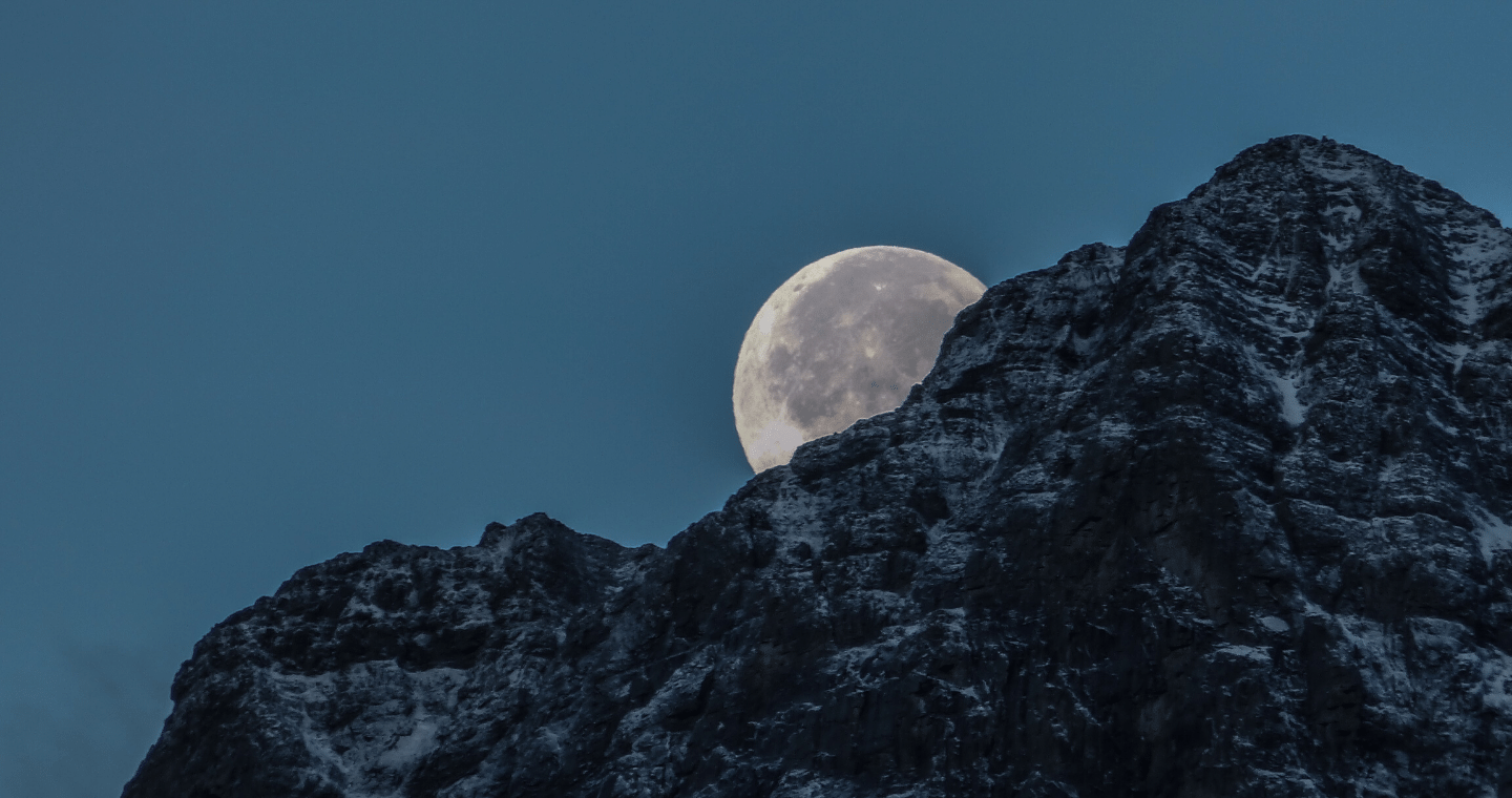 A full moon against a dark blue night sky is half-obscured by a mountain. Photo by Kym MacKinnon on Unsplash.