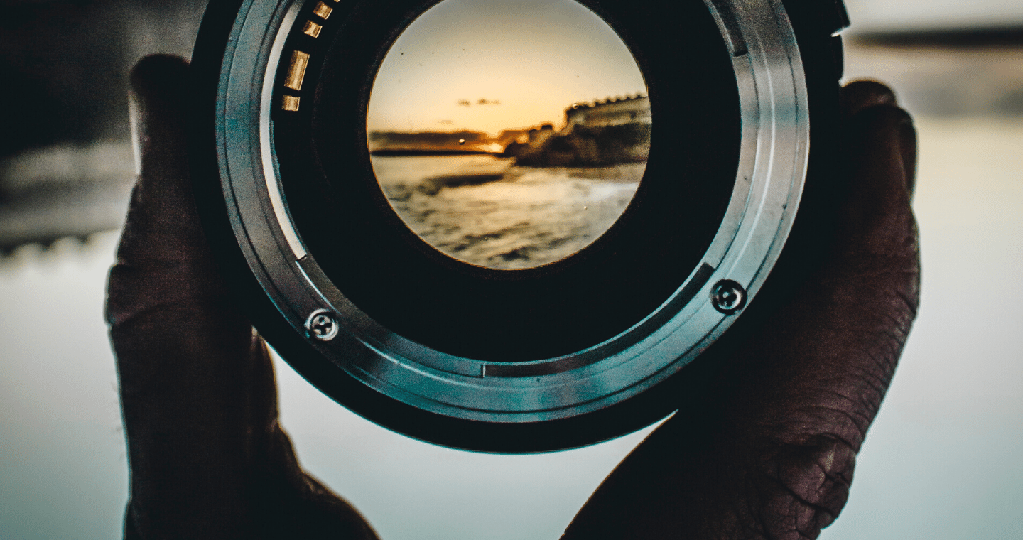 A close-up view of a person's hand holding a camera lens through which a rocky beach view is visible.