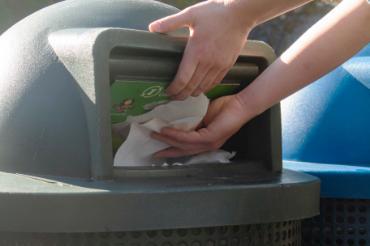 Someone throwing a paper towel into a green compost bin indicated by a compost symbol.