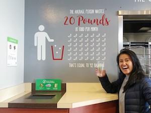 happy student holding a compostable cup above compost bin in c street