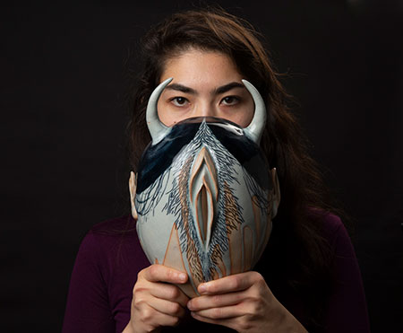 a photographic portrait of a person with dark hair holding a sculpted spirit in front of the lower part of her face