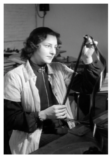 A black and white photo of a white woman holding up a long film strip vertically between her hands and looks intently at it.