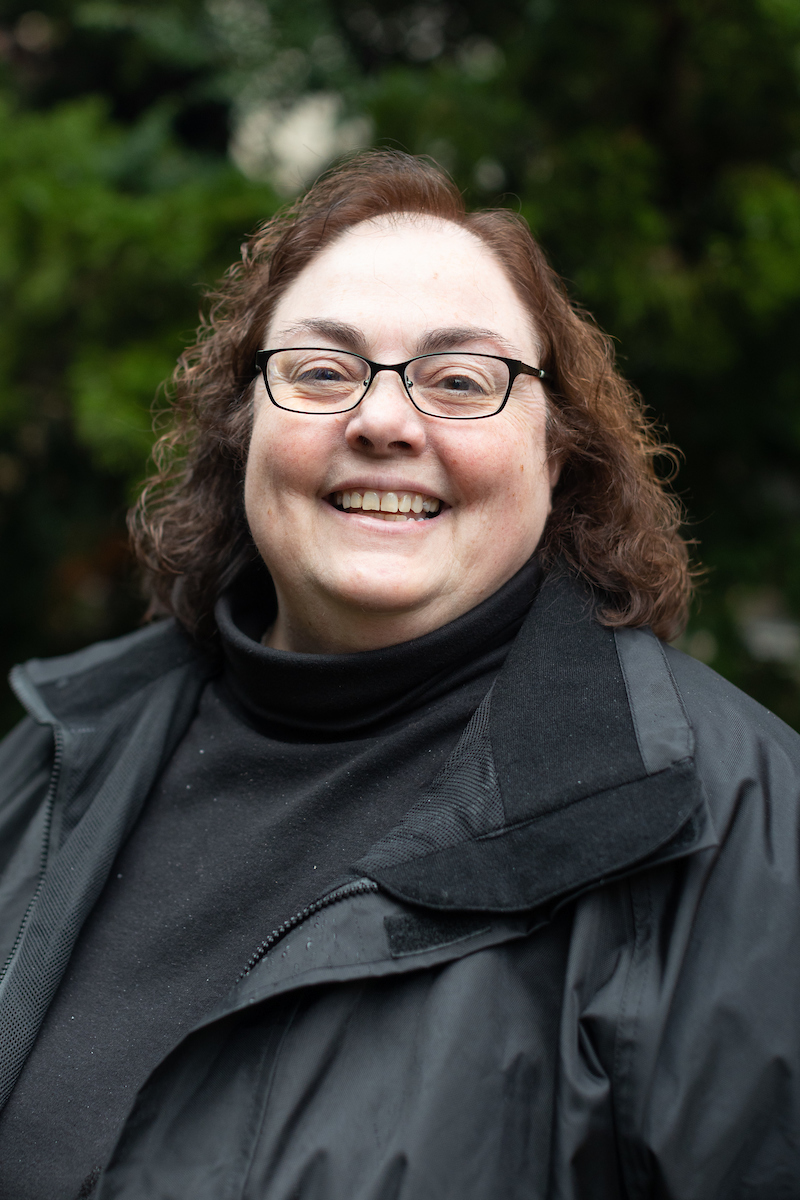 Kim smiling, curly shoulder length hair, glasses, black shirt and jacket