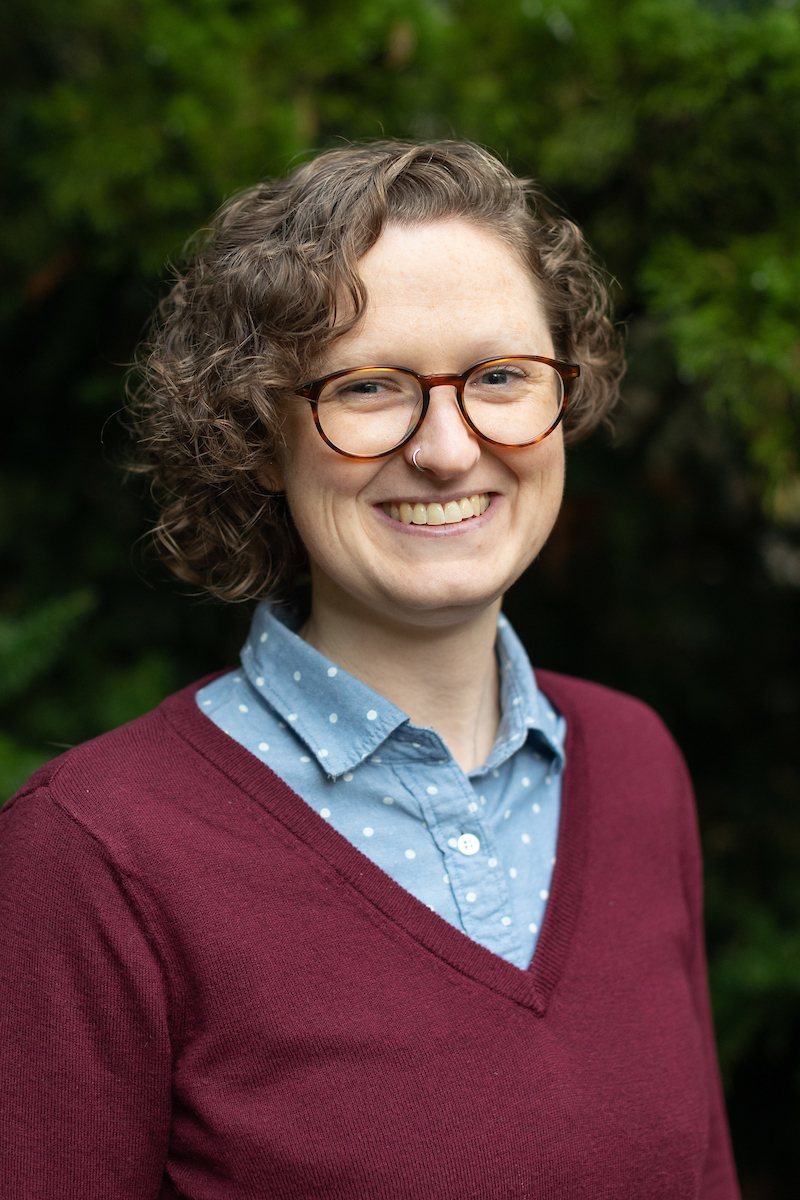 Maggie smiling, curly chin length hair, glasses, blue shirt under v neck sweater