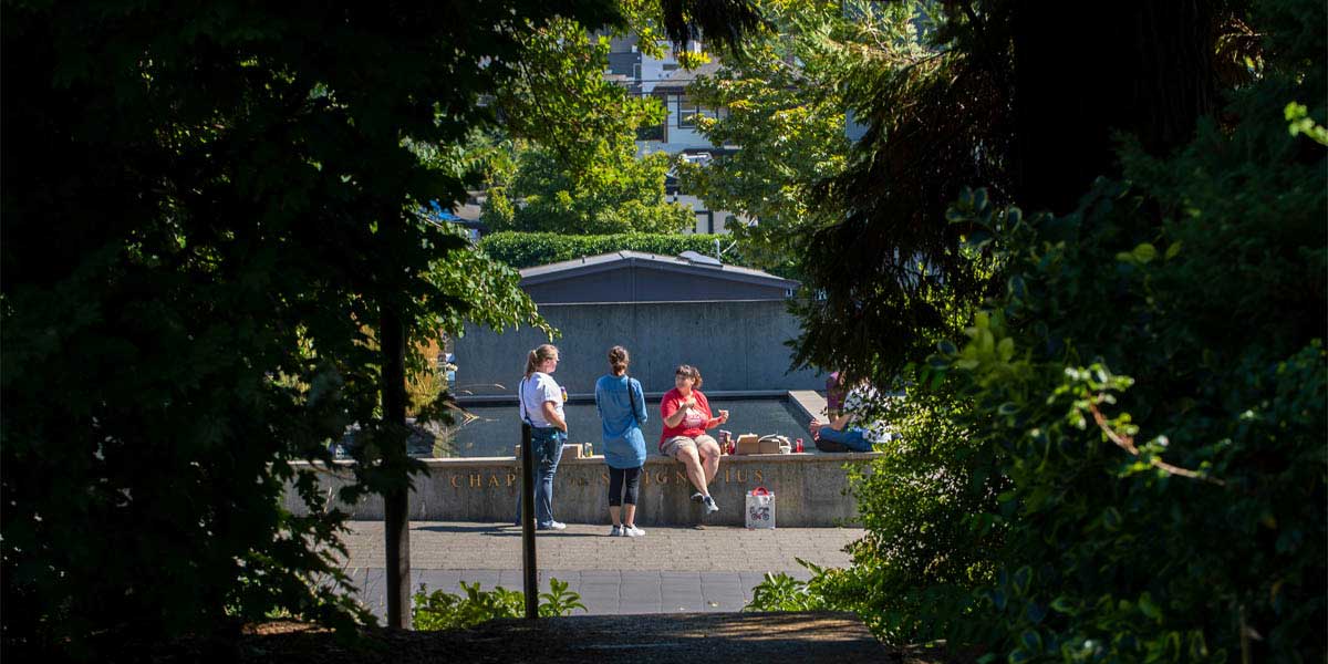 A campus scene taken from a distance. A small group of students sitting outside and surrounded by campus greenery.