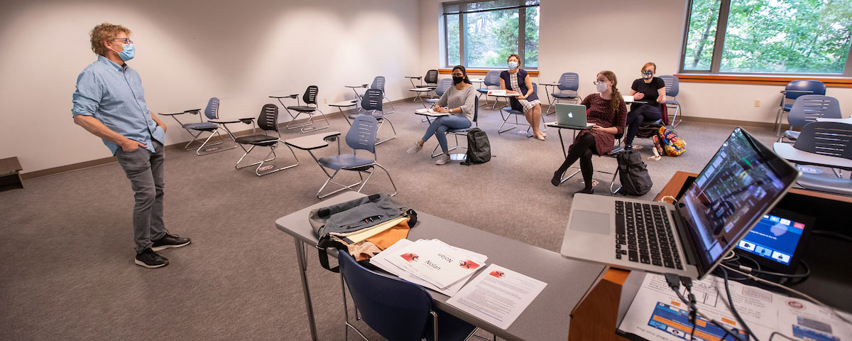 Professor Kevin Lundeen, MS, lectures in a classroom of 4 students in-person and more students online as shown on a nearby laptop