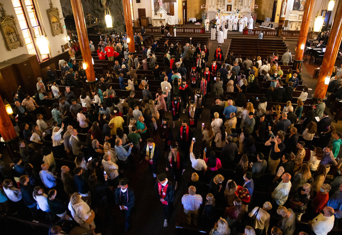 Attendees standing in church pews while graduates wearing caps and gowns walk away from the altar