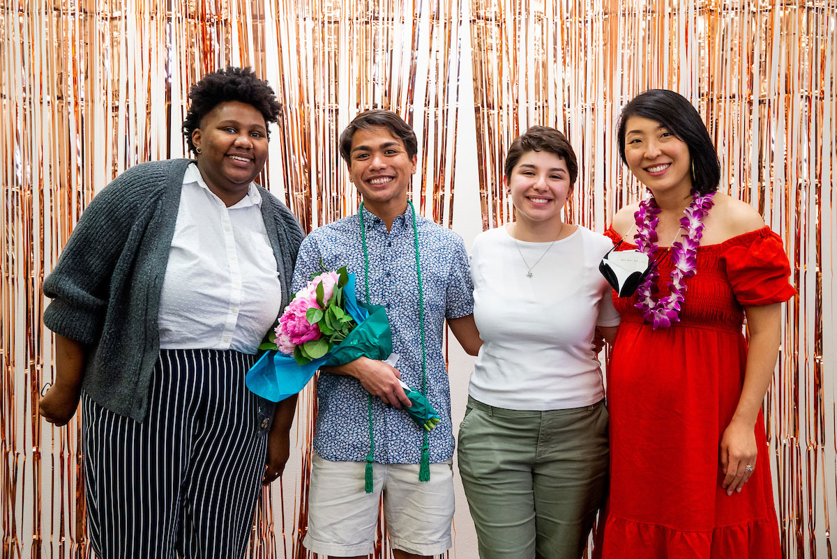 Two graduates pose in between two Office of Multicultural staff members with metallic streamers in the background