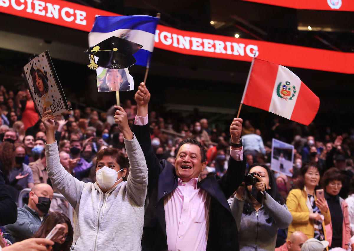 Excited family and friends in the audience cheer their grads on with flags and homemade signs