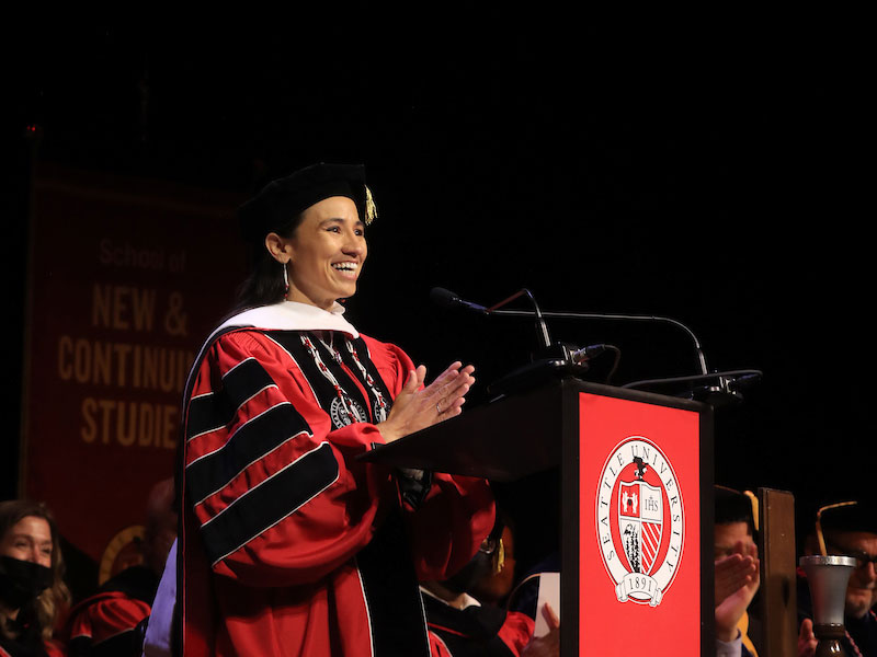 U.S. Representative Sharice Davids standing on stage behind a podium dressed in graduation regalia.