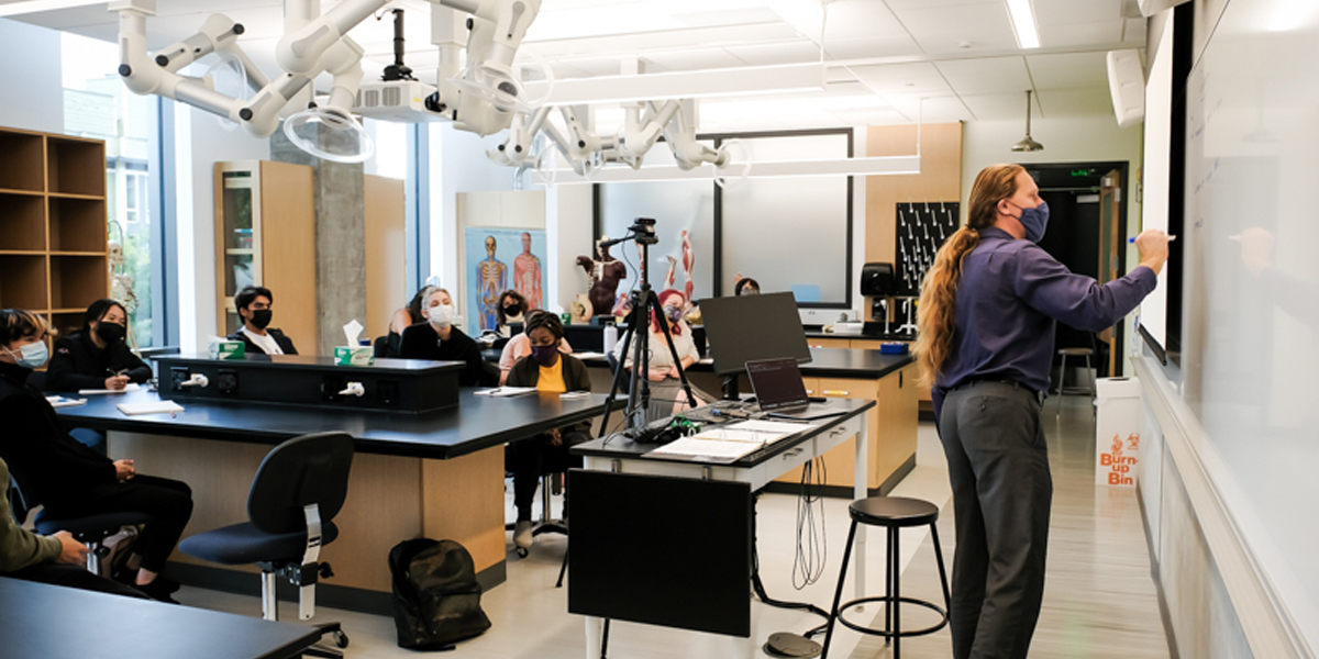 Photo of students in masks sitting in classroom led by professor writing on board.