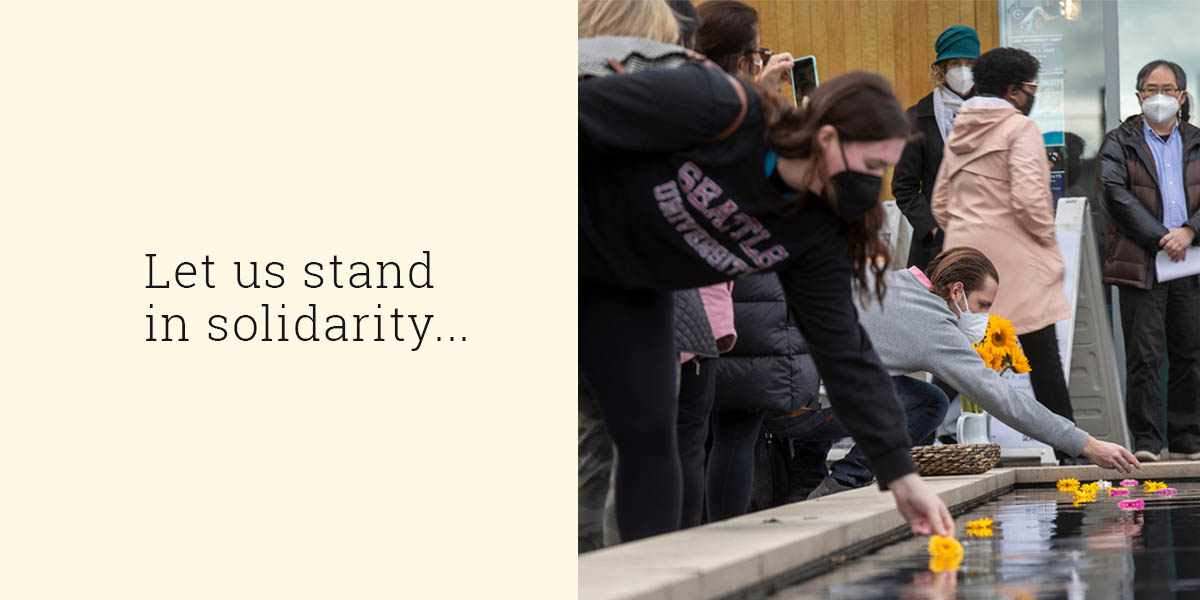 Campus community members lay flowers in the reflection pond at the vigil. Text reads: Let us stand in solidarity.