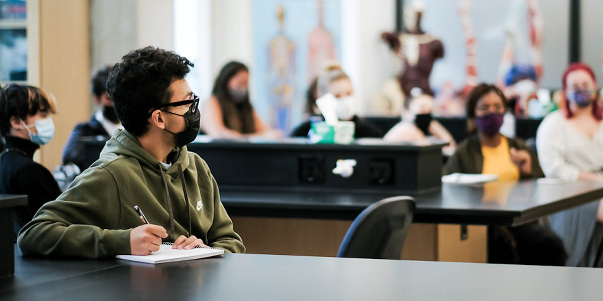 Masked students in a science class. Student closest to camera sits with an open notebook while holding a pen ready to write notes.