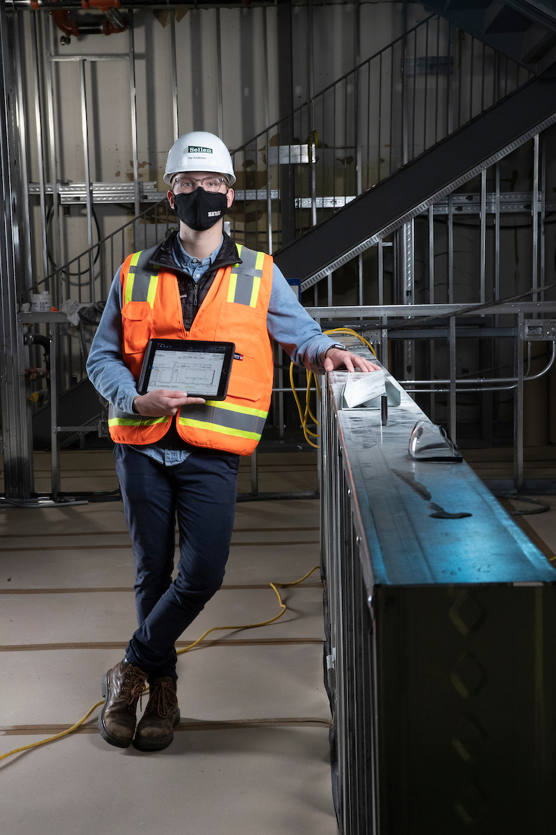 Per Andersen, ’21, wearing a Sellen construction hat and an orange safety vest, poses with a tablet displaying blueprints.