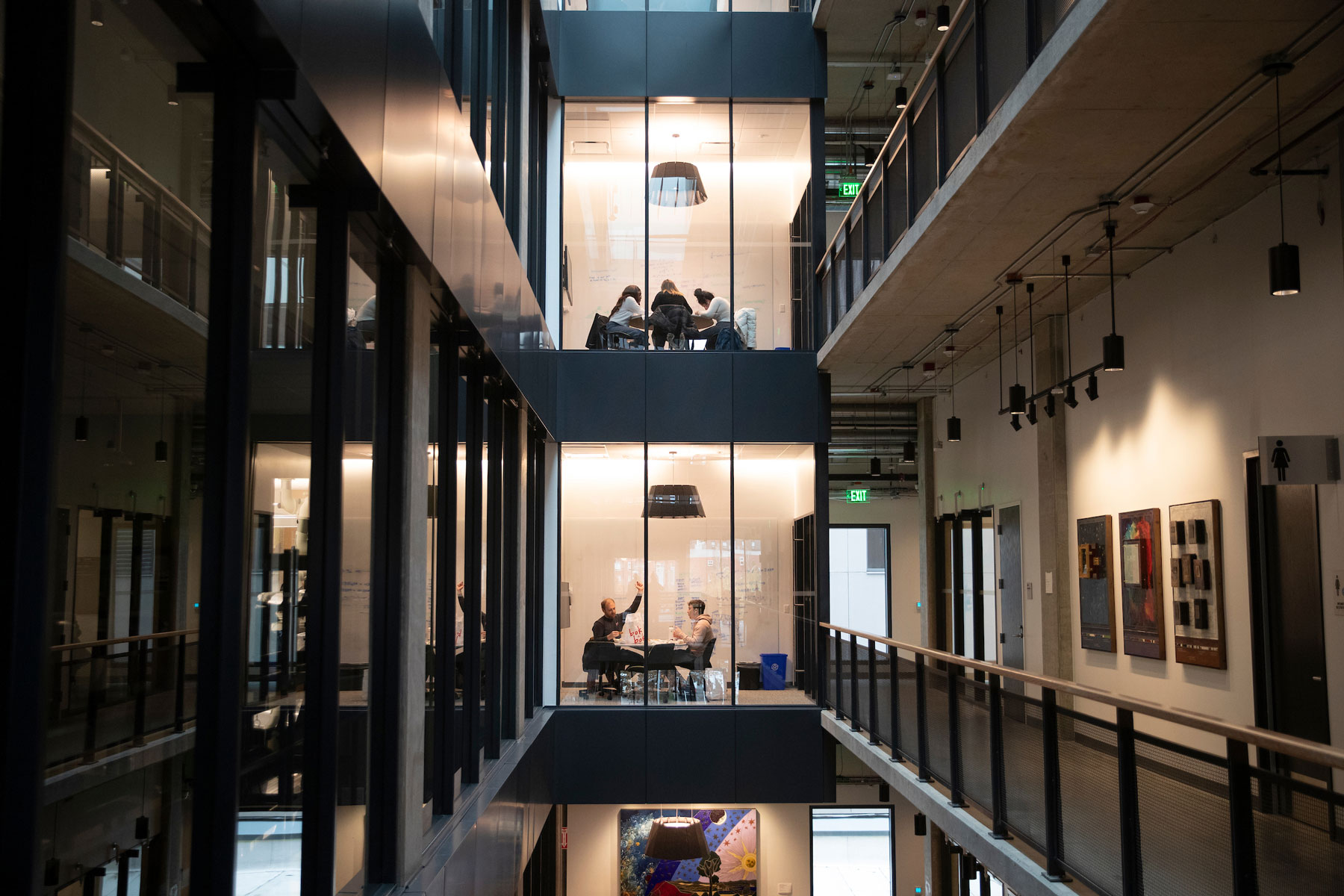 A multi-story interior view of a sleek new building filled with groups studying in rooms with glass doors and pendant lights